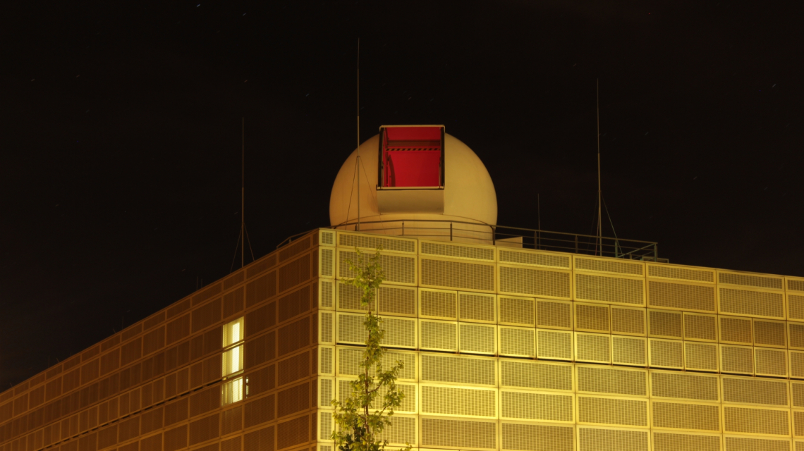 Das golden leuchtende Gebäude der Matematisch-Naturwissenschaftlichen Fakultät der Universität Potsdam bei Nacht, oben auf dem Dach ist die Teleskopkuppel mit geöffnetem Kuppelspalt zu sehen.
